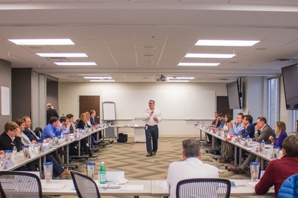 a man is giving a presentation in a conference room where people are seated at tables arranged around him