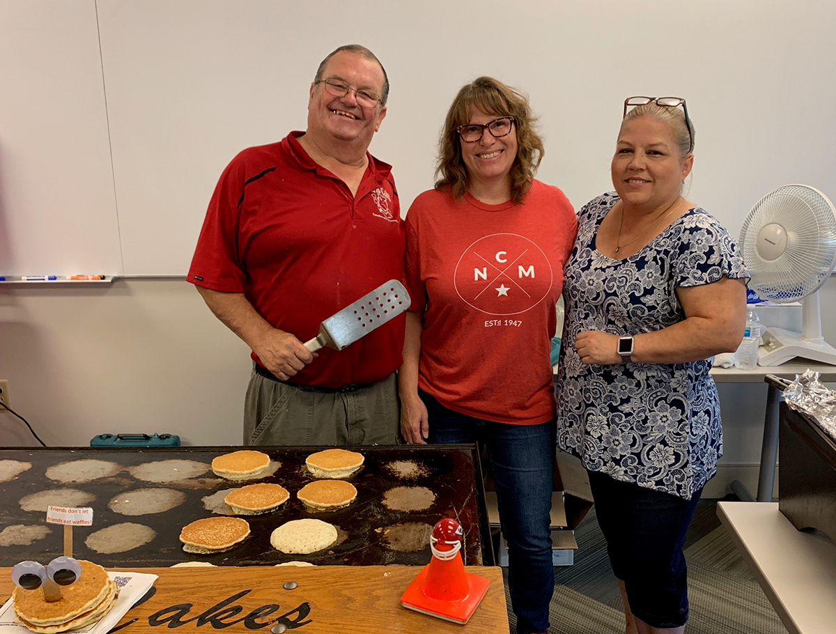 3 people standing and smiling behind a griddle where pancakes are frying