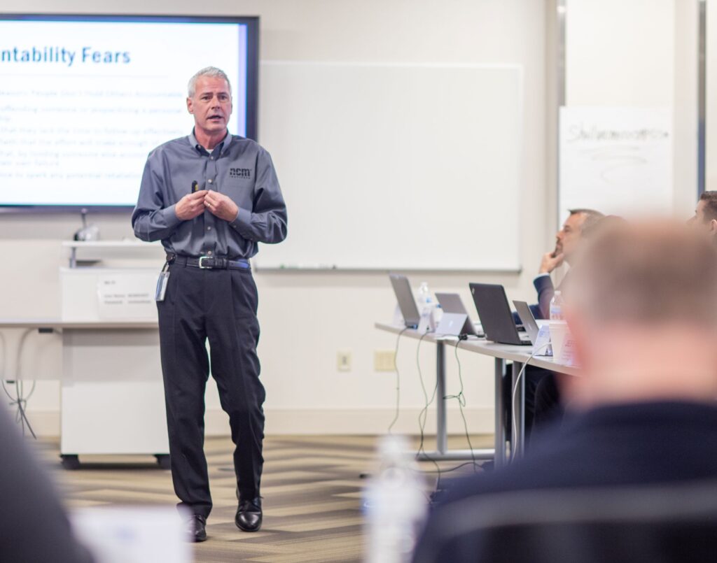 A man in an NCM uniform is standing giving a presentation in the middle of classroom during an automotive training session.