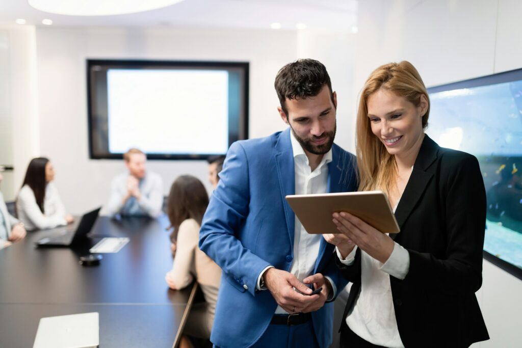 a man and a woman look at a tablet she is holding between them in a conference room setting