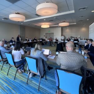 a group of people is gathered around four tables arranged as a square in a conference room for an NCM training session.