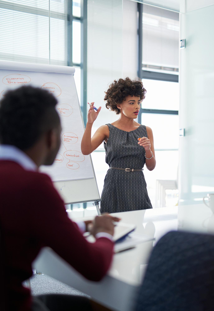 a business woman is standing next to a board and presenting to a group of people.