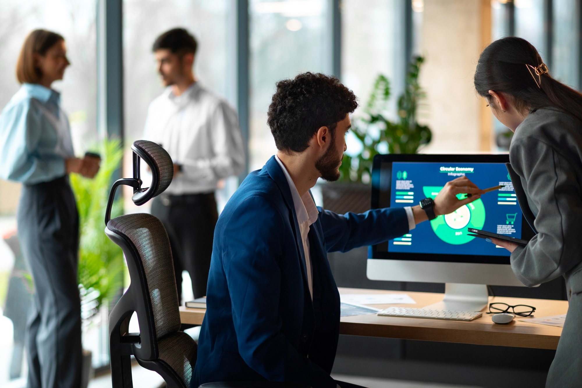 two people discuss a graph on a desk top computer in an office setting. they are facing away from the camera.