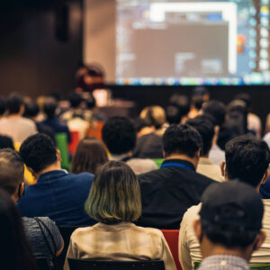 Image is from behind an audience viewing a powerpoint presentation at a dealer meeting. The screen is blurred out, only the backs of heads are visible.