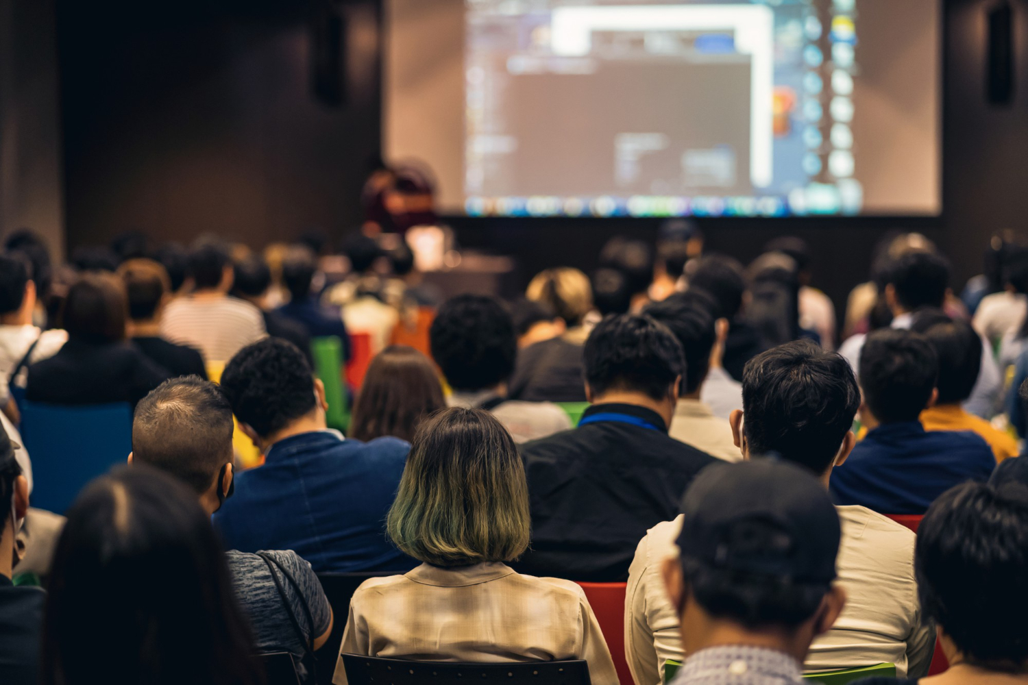 Image is from behind an audience viewing a powerpoint presentation at a dealer meeting. The screen is blurred out, only the backs of heads are visible.