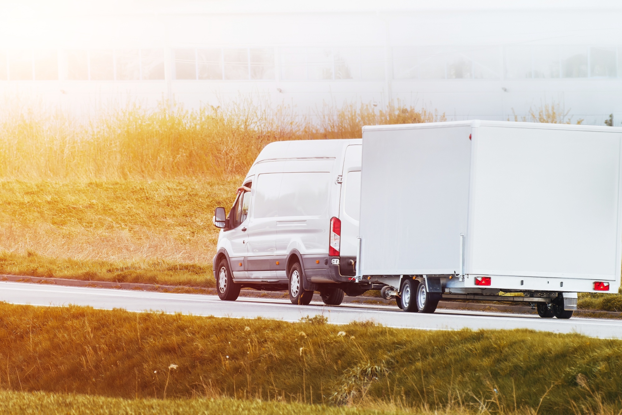 a white sprinter van is pulling a white cargo trailer about the same size as the van on a pave road cutting through a grass landscape