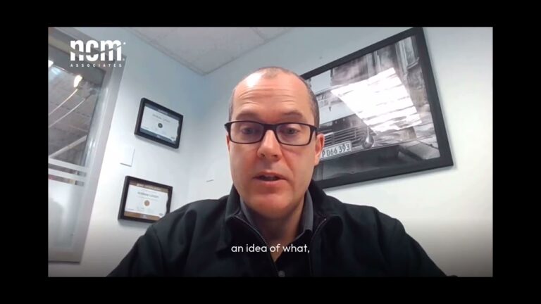 Andrew Linton is wearing glasses and a black jacket sitting in an office setting during an online coaching session.