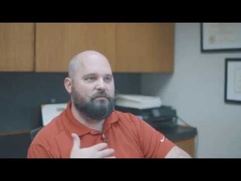 Chris Thomas, a man with a shaved head and a full beard, wearing a red polo shirt and sitting in an office setting