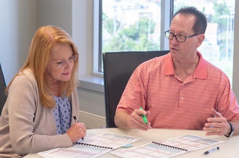 A woman and a man sit at a table looking at spiral bounding spreadsheets with writing utensils in hand.