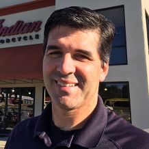 a headshot of man in a blue collared shirt smiling and facing the camera.