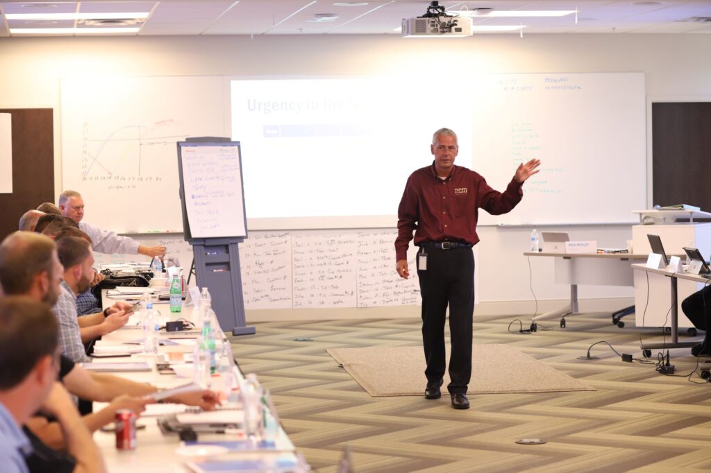 A man in an NCM uniform stands in the middle of a classroom giving a presentation during a training session. On the left hand side, adult students sit at tables taking notes and paying attention.