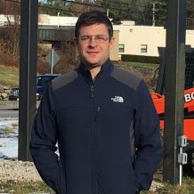 An man in glasses and a blue jacket is standing facing the camera and smiling.