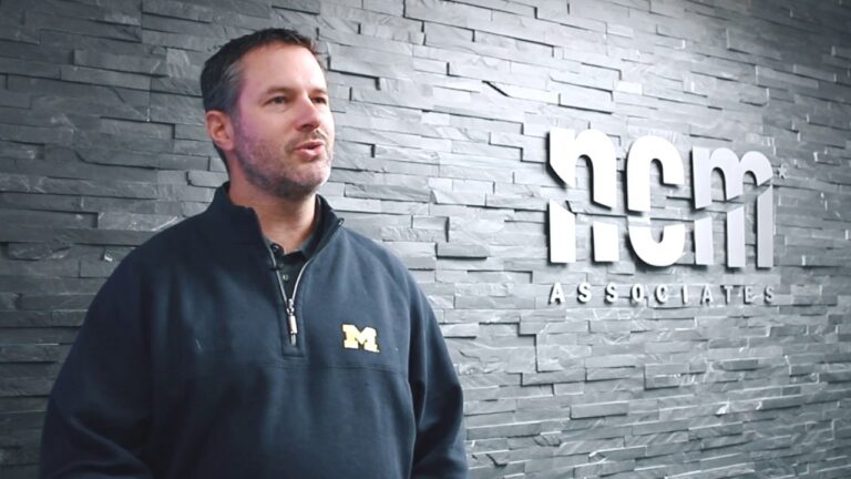 Joe Zimmerman, a man in a black quarter zip pullover, stands in front of a wall with the NCM Associates logo in the background