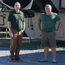 two men in green polos and brown pants are standing in front of a RV trailer.