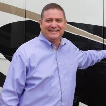Mike Regain stands in front of an RV, smiling and facing the camera. He's wearing a purple collared shirt.
