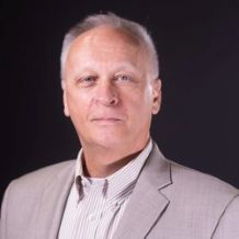 headshot of Mike Udell from mid chest up, a white man with gray hair in a gray suit.