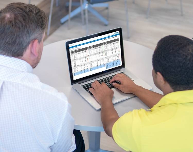 two men are looking at a laptop while one of them types on it. There are spreadsheets displayed on the screen