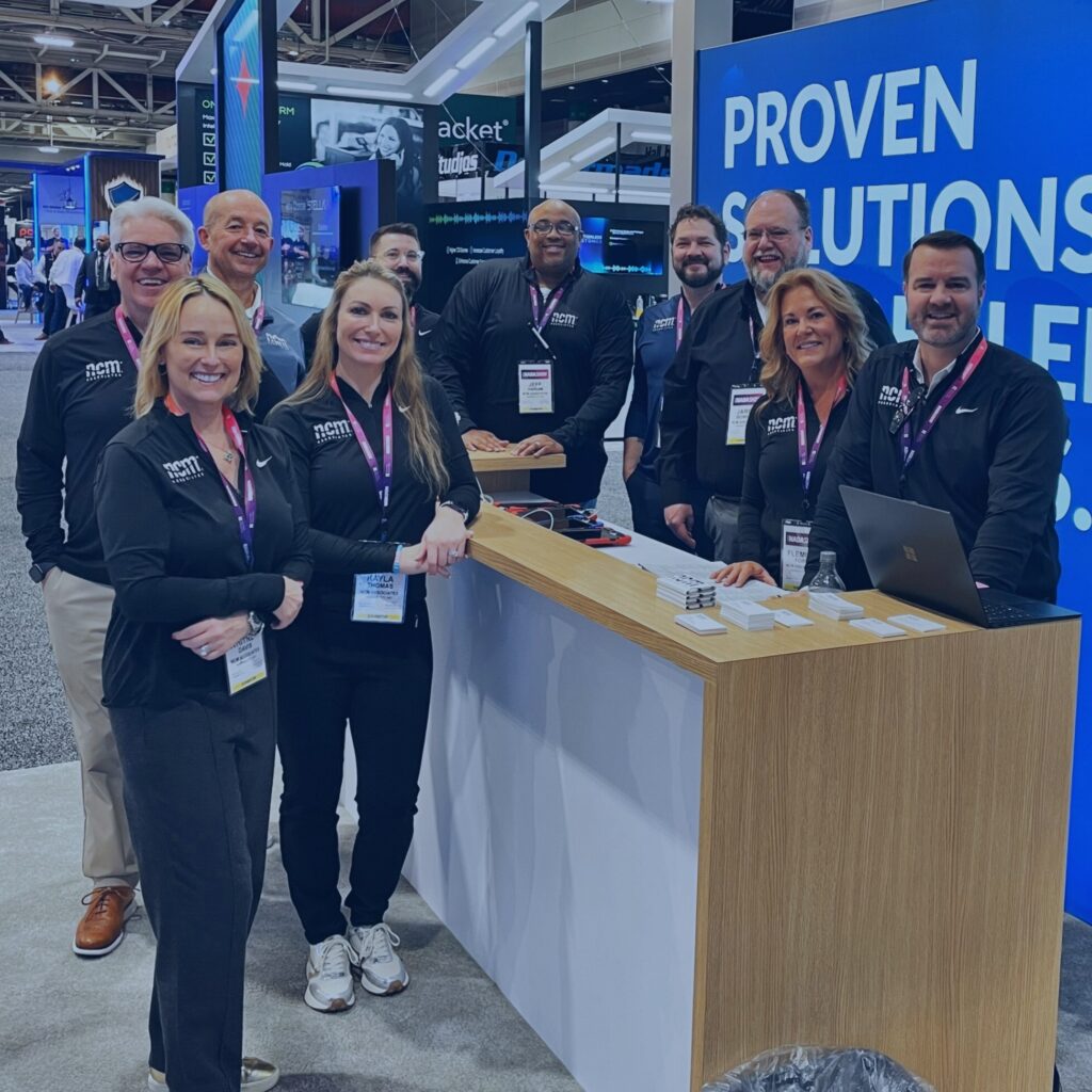 A portion of the NCM team is standing in their booth at a trade show. they are all wearing the NCM uniform and smiling.