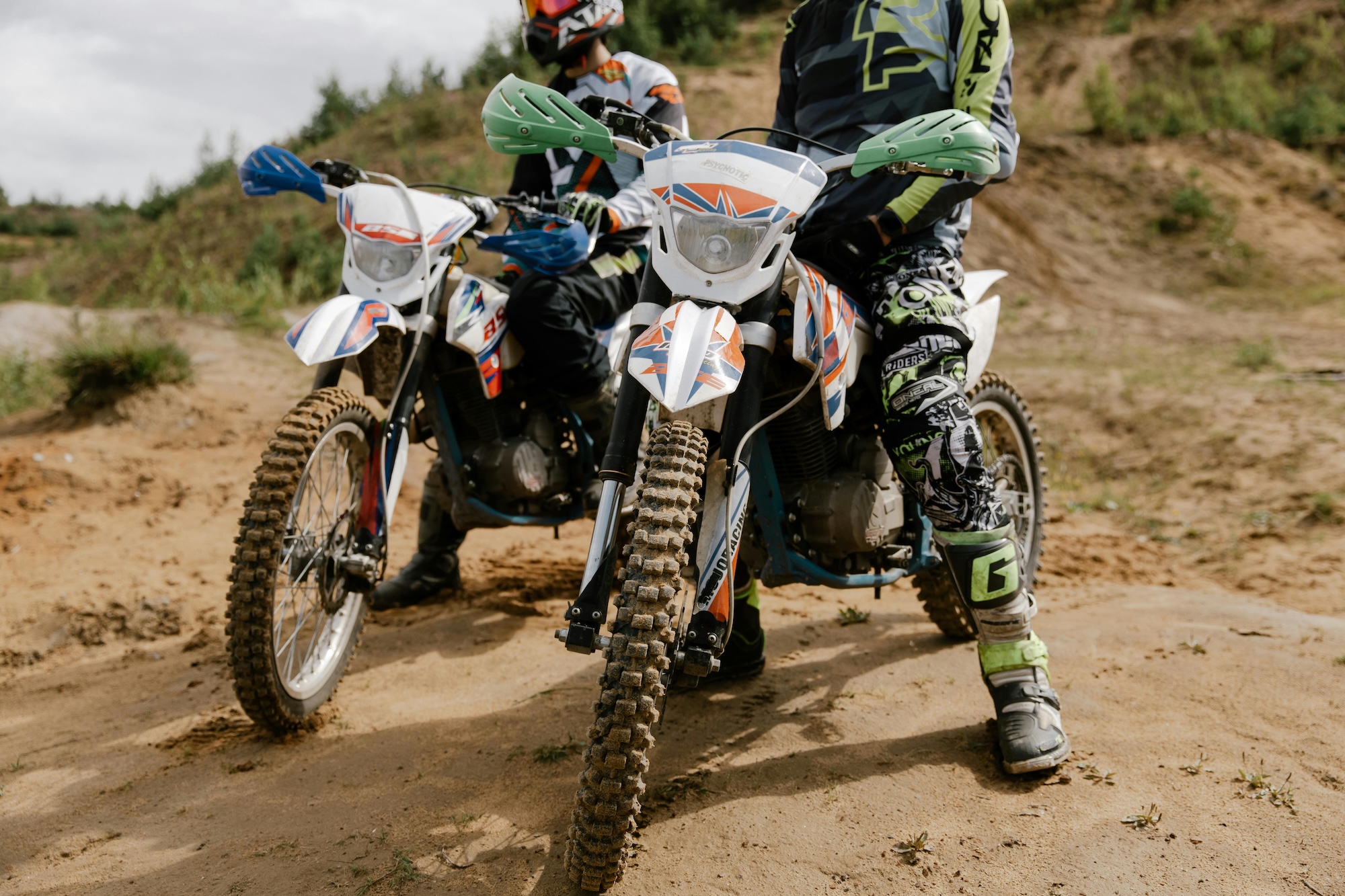 Two people are sitting on dirt bikes on a sandy hill