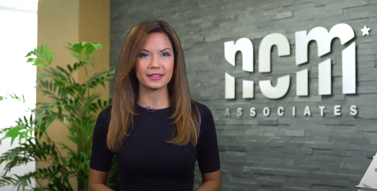 A woman with long brown hair and a black shirt smiles in front of NCM logo