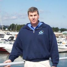Rob Soucy stands on a pier wearing a blue jack and facing the camera. There are boats docked in the background.