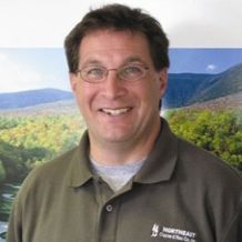 Scott Riccio is smiling and facing the camera with a landscape in the background. He is wearing glasses and a green shirt.