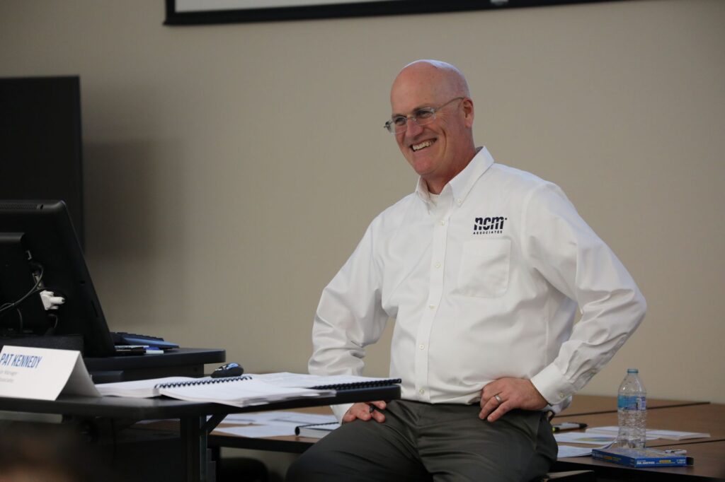 A man in a white NCM collared shirt sits at the front of a classroom smiling during a training session.