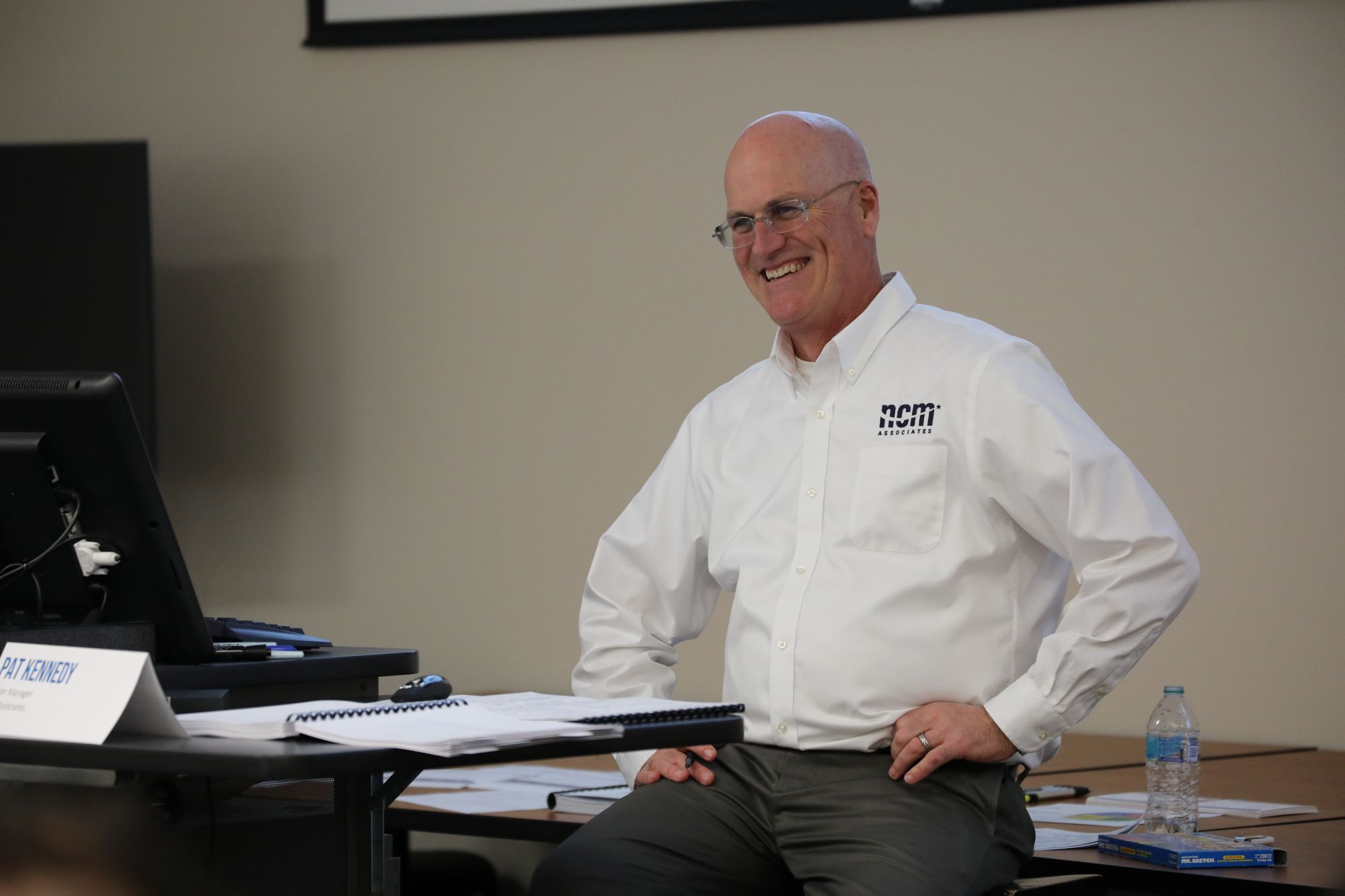 A man in a white NCM collared shirt sits at the front of a classroom smiling during a training session.