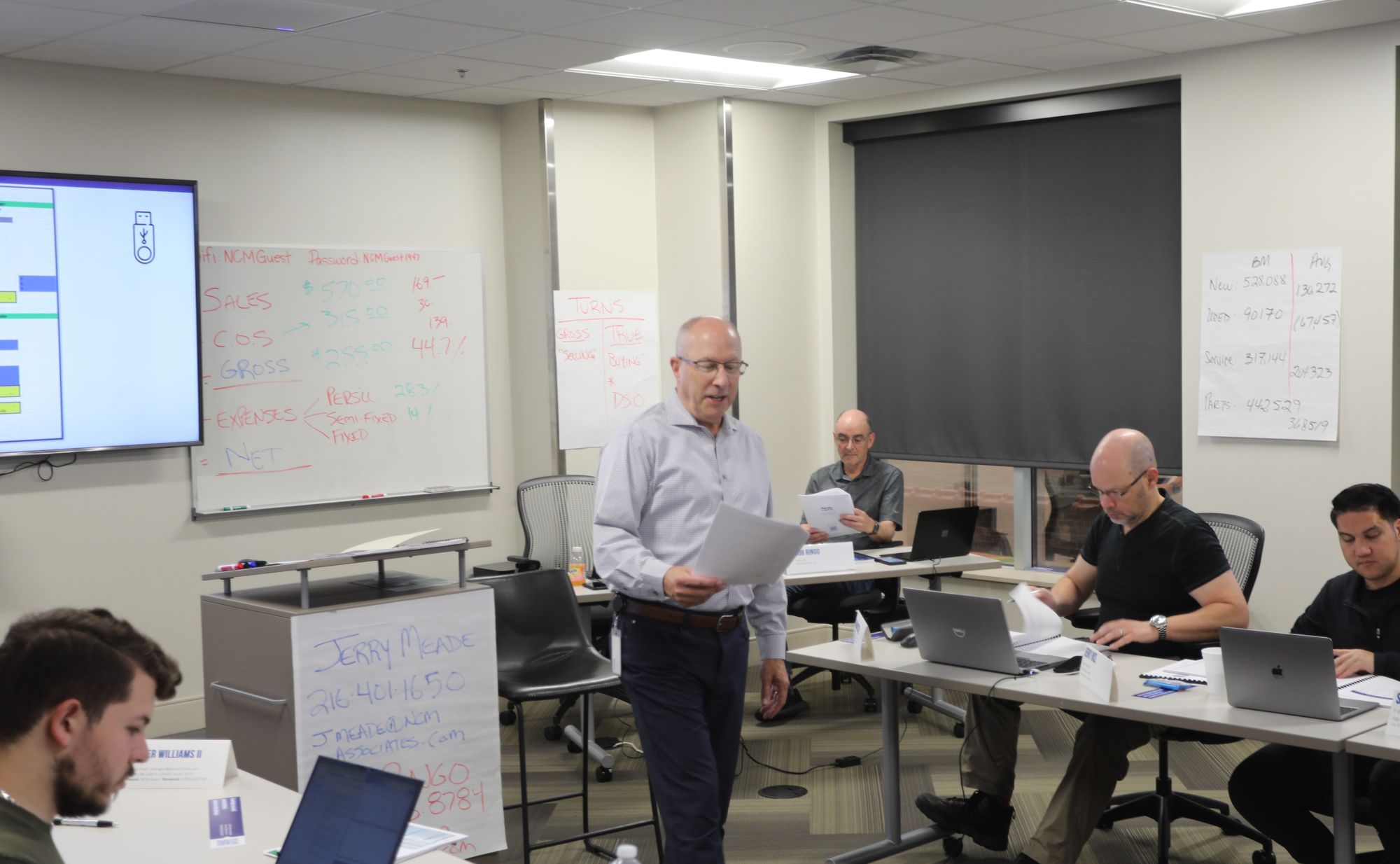 A man reads from a paper in his hand. He is standing in the middle of a classroom. Adult students sit at desks also looking at their papers to follow along.