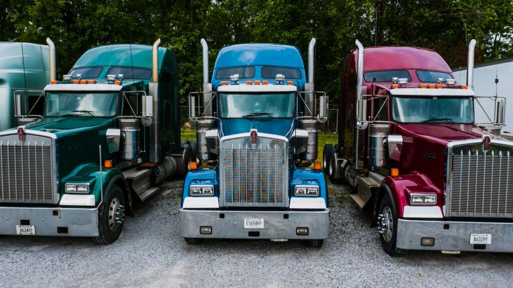 3 semi trucks lined up next to each other. one is green, one is blue, one is pink.