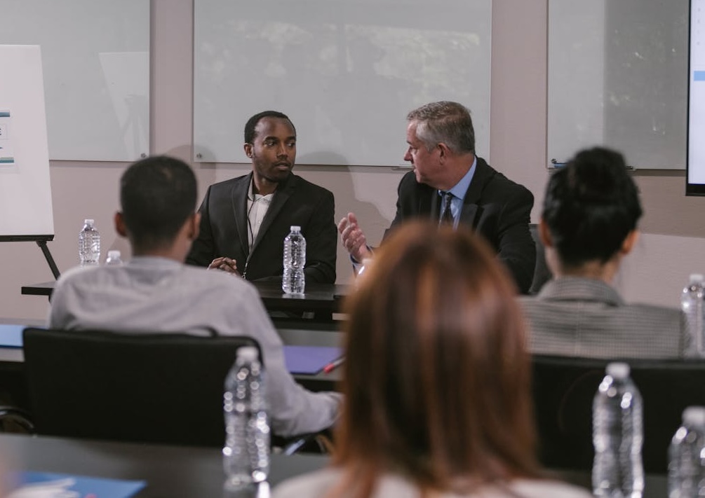 two men sit at the front of a group of people discussing something